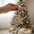 Hand holding a Christmas ornament with a house design and 'Our Best Year in our New Home' text, blurred Christmas tree in the background.